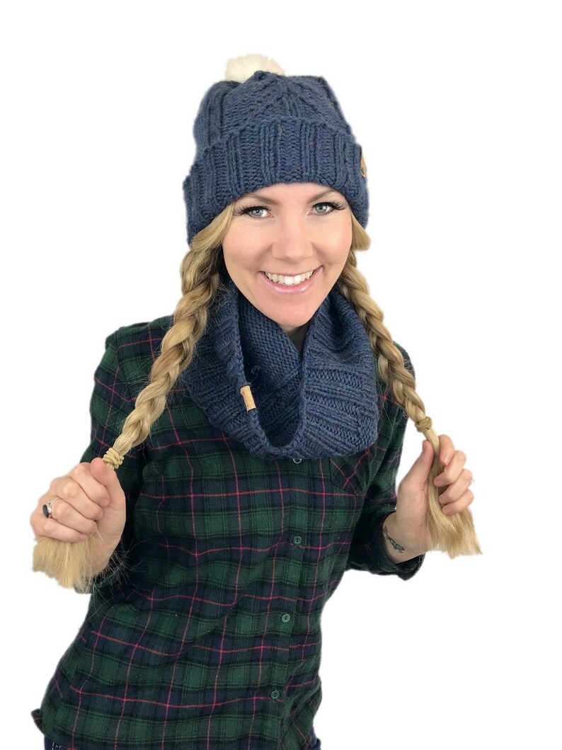 May include: A woman wearing a blue knit hat with a white pom pom and a matching blue knit scarf. She is wearing a green and black plaid shirt and has her hair in two braids.