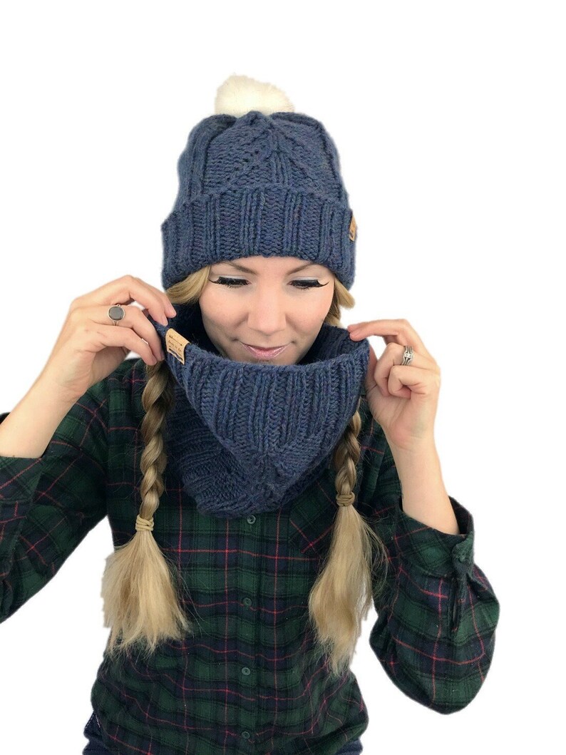 May include: A blue knitted hat with a white pom pom and a matching blue knitted neck warmer. The hat and neck warmer have a cable knit pattern.