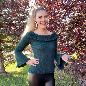 May include: A dark green knitted sweater with a wide, off-the-shoulder neckline and bell sleeves. The sweater is made of a soft, textured yarn.
