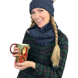 May include: A woman wearing a blue knit hat with a white pom pom, a matching blue knit scarf, and a green and black plaid shirt is holding a tall ceramic mug with a Christmas scene featuring Santa Claus.