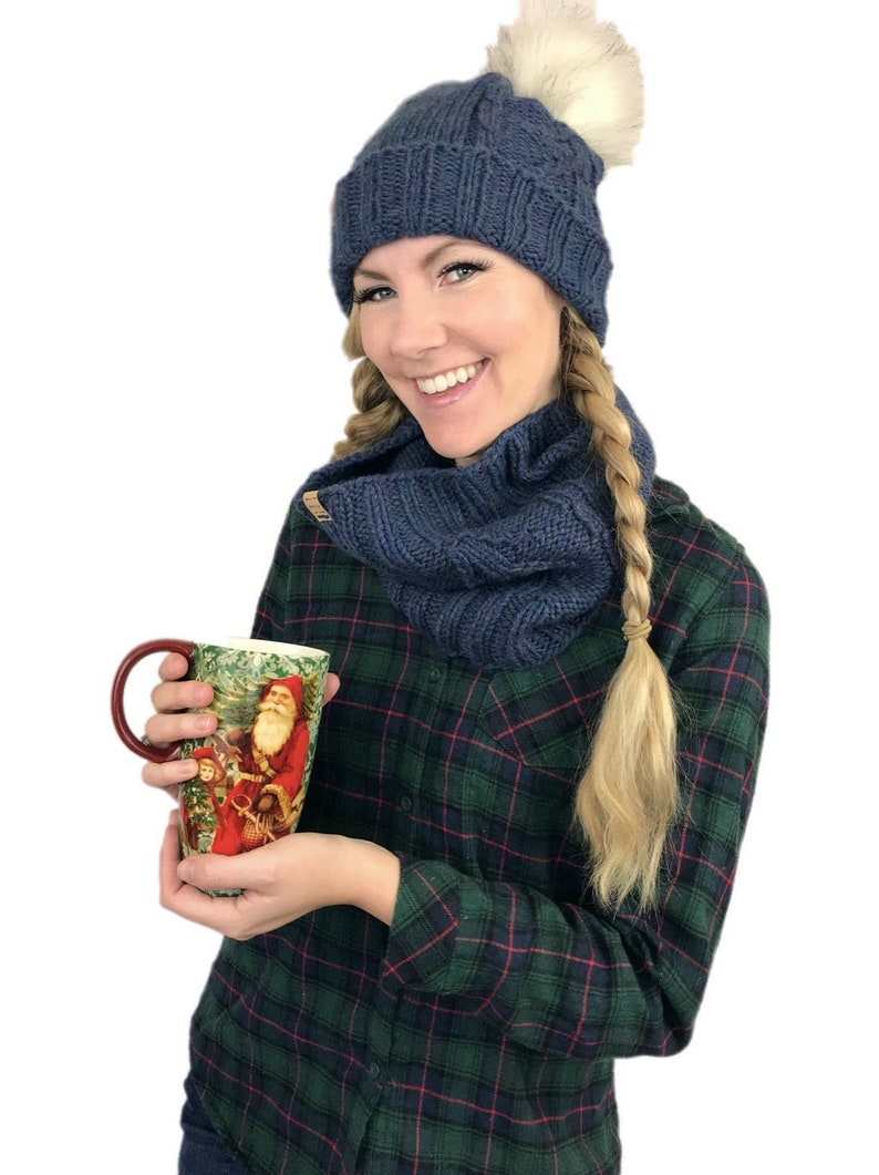 May include: A woman wearing a blue knit hat with a white pom pom, a matching blue knit scarf, and a green and black plaid shirt is holding a tall ceramic mug with a Christmas scene featuring Santa Claus.