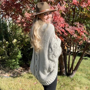 May include: A woman wearing a gray cable knit sweater with a brown fedora hat. She is standing in front of a tree with red leaves.