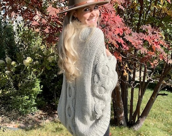 May include: A woman wearing a grey cable knit sweater with a brown fedora hat. She is standing in front of a tree with red leaves.