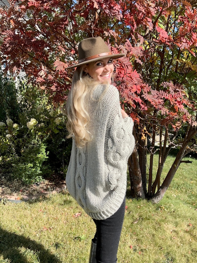 May include: A woman wearing a gray cable knit sweater with a brown fedora hat. She is standing in front of a tree with red leaves.