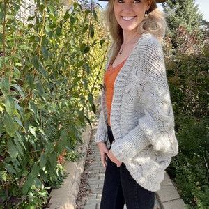 May include: A woman wearing a light gray cable knit cardigan with a brown fedora hat, a burnt orange tank top, and black jeans.