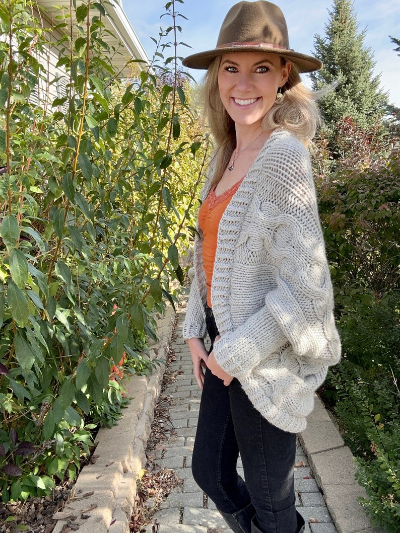 May include: A woman wearing a light gray cable knit cardigan with a brown fedora hat, a burnt orange tank top, and black jeans.