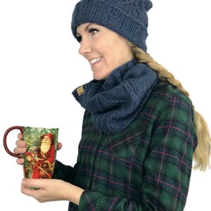 May include: A woman wearing a blue knitted hat with a white pom pom and a matching blue knitted scarf. She is holding a red mug with a Christmas scene on it.