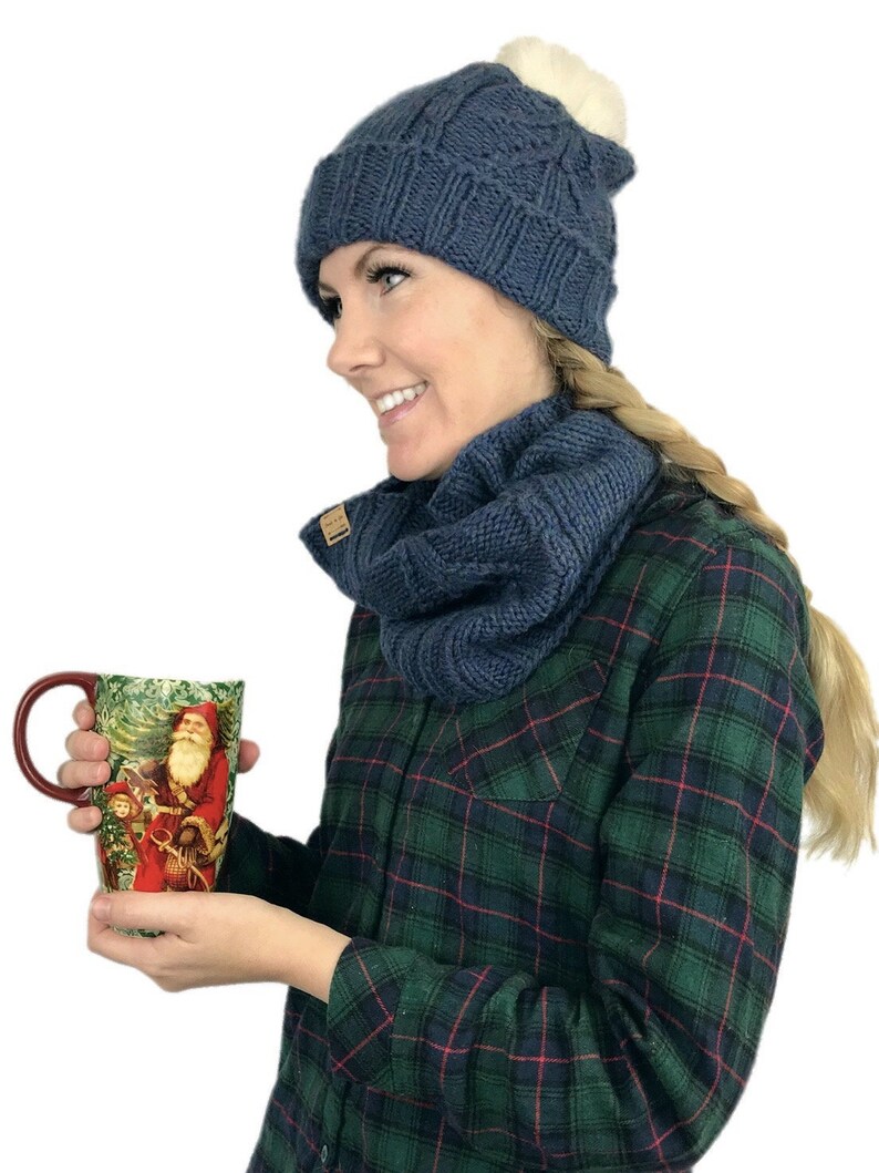 May include: A woman wearing a blue knitted hat with a white pom pom and a matching blue knitted scarf. She is holding a red mug with a Christmas scene on it.