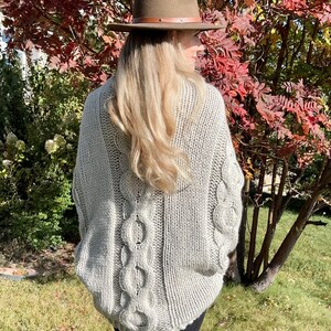 May include: A gray cable knit sweater with a loose fit and a wide neckline. The sweater has a cable knit pattern on the back and a plain knit pattern on the front. The sweater is being worn by a person with long blonde hair and a brown hat.