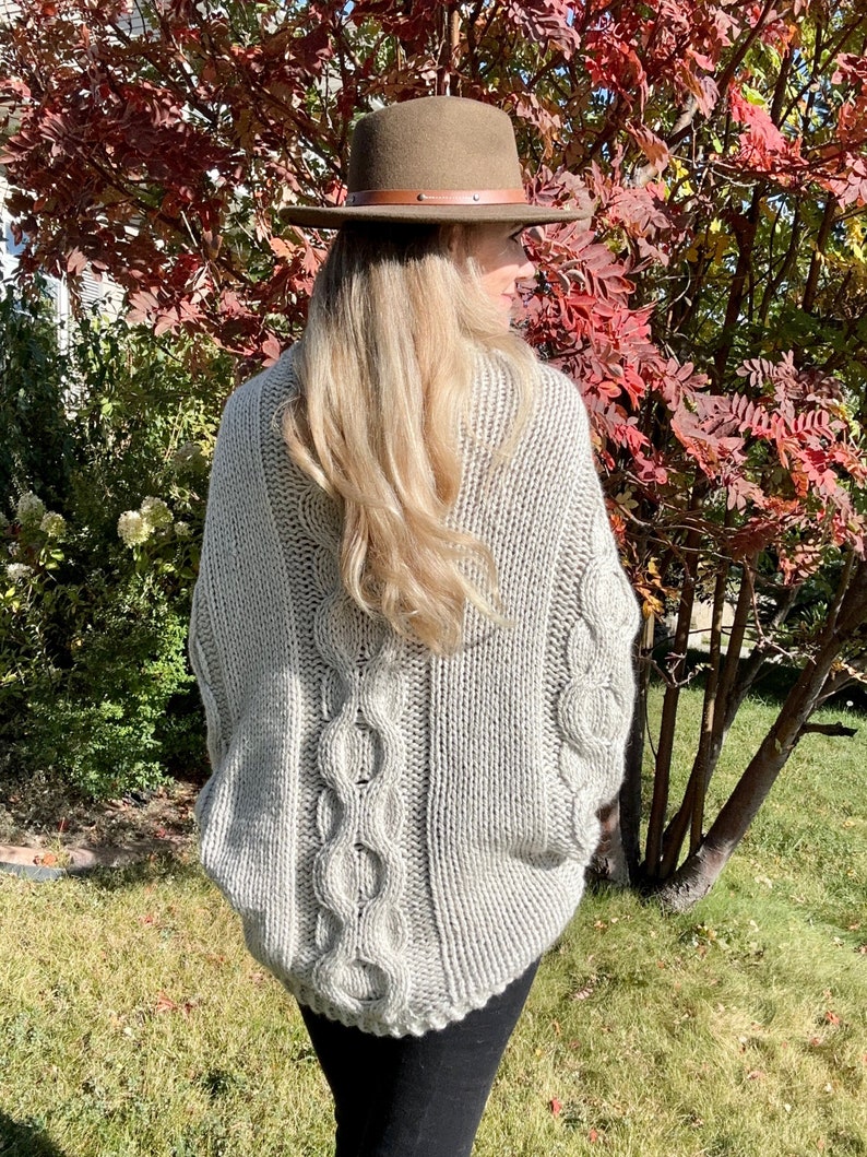 May include: A gray cable knit sweater with a loose fit and a wide neckline. The sweater has a cable knit pattern on the back and a plain knit pattern on the front. The sweater is being worn by a person with long blonde hair and a brown hat.