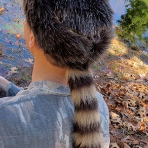 May include: A brown and black fur hat with a raccoon tail attached. The hat is being worn by a person with a camouflage shirt.