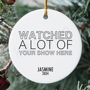 May include: White round Christmas ornament with black text that reads "Watched a lot of your show here" and "Jasmine 2024". The ornament is hanging on a green Christmas tree.
