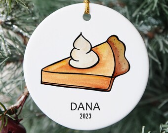 Peut inclure: Une boule de Noël en céramique blanche avec une illustration dessinée à la main d'une part de tarte à la citrouille avec de la crème fouettée. La boule est décorée du nom "DANA" et de l'année "2023".