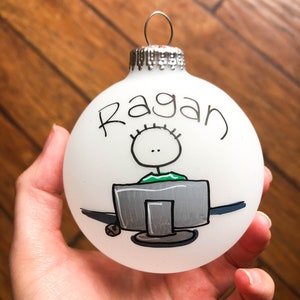 May include: White Christmas ornament with a hand-drawn illustration of a person sitting at a computer. The ornament is personalized with the name "Ragan".