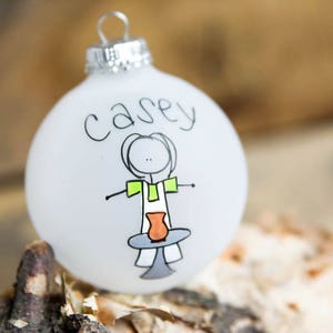 May include: White frosted glass Christmas ornament with the name "Casey" written in black marker and a stick figure drawing of a person holding a pottery piece.