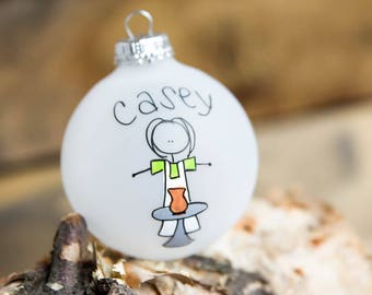 May include: White frosted glass Christmas bauble with the name "Casey" written in black marker and a stick figure drawing of a person holding a pottery piece.