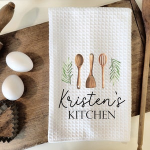 Könnte beinhalten: Ein weißes Küchentuch mit Waffelstruktur. Das Tuch zeigt eine Aquarell-Illustration von drei Küchenutensilien aus Holz und grünen Zweigen. Der Text "Kristen's Kitchen" ist in Schwarz unter der Illustration gedruckt.