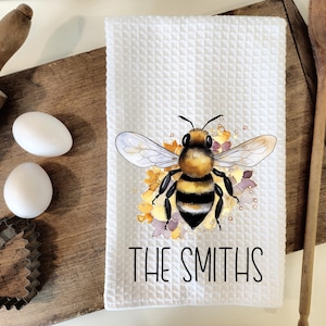 Può includere: Un asciugamano da cucina bianco con un'illustrazione ad acquerello di un'ape e fiori. Il testo "THE SMITHS" è stampato in nero sotto l'ape.