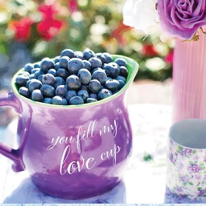 May include: A purple jug filled with blueberries, with the words "you fill my love cup" written on the side. The jug is sitting on a white table with a floral patterned mug and a vase of purple flowers in the background.