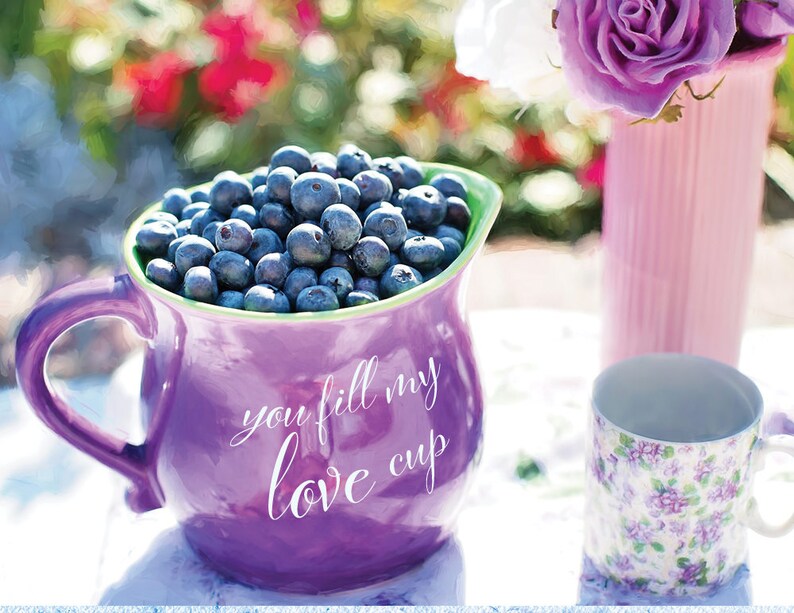 May include: A purple jug filled with blueberries, with the words "you fill my love cup" written on the side. The jug is sitting on a white table with a floral patterned mug and a vase of purple flowers in the background.
