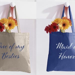 May include: Two tote bags with flowers. One bag is beige with blue text that says "One of my Besties". The other bag is blue with white text that says "Maid of Honour".