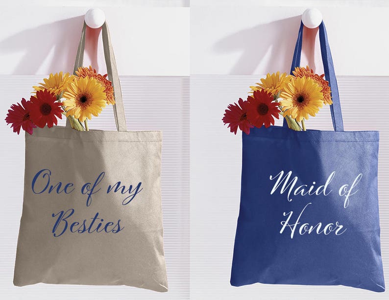 May include: Two tote bags with flowers. One bag is beige with blue text that says "One of my Besties". The other bag is blue with white text that says "Maid of Honour".