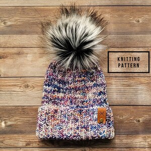 May include: A colourful knitted beanie with a white and black faux fur pom pom. The beanie has a fold-over cuff and a brown leather patch with a stitched X.