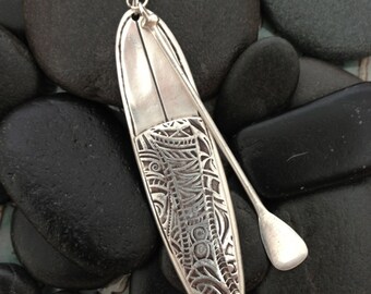May include: A silver pendant necklace featuring a detailed design of a surfboard and paddle. The pendant is hanging from a silver chain.