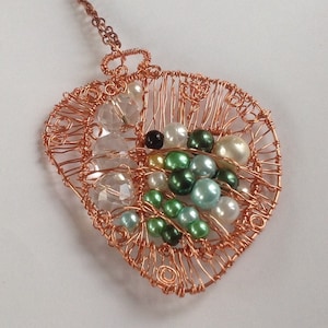 May include: A copper wire pendant necklace with a heart-shaped design. The pendant is filled with various colored beads, including white, green, and black. Clear crystal-like beads are also incorporated. The necklace chain is also copper-colored.