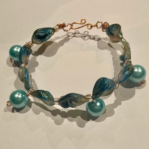 May include: A handcrafted bracelet with iridescent blue and white shell-shaped beads and light blue pearl beads. The bracelet is strung on copper wire with a copper hook clasp. The jewellery is designed for a delicate and elegant look.
