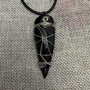 May include: A black obsidian arrowhead pendant, secured with silver-coloured wire, hanging from a black cord necklace. The arrowhead is a deep, glossy black, and the wire is intricately wrapped around it. The necklace is displayed against a textured grey background.
