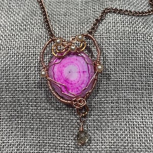 May include: A handcrafted necklace featuring a pink and white agate pendant. The pendant is wire-wrapped in copper-coloured metal and accented with small pearls. A small crystal hangs from the bottom of the pendant.