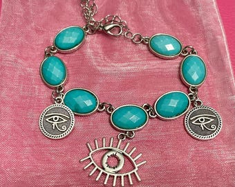 May include: Silver-toned bracelet featuring oval turquoise-colored stones and dangling charms. The charms include two round pendants with an eye design and a larger open eye charm. The bracelet is displayed on a pink surface.