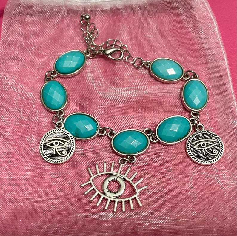 May include: Silver-toned bracelet featuring oval turquoise-colored stones and dangling charms. The charms include two round pendants with an eye design and a larger open eye charm. The bracelet is displayed on a pink surface.