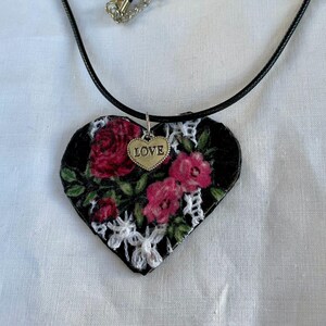 May include: A heart-shaped pendant necklace with a black cord. The pendant features a floral design with red roses and white lace on a black background. A small silver heart charm with the word "LOVE" hangs below the pendant.