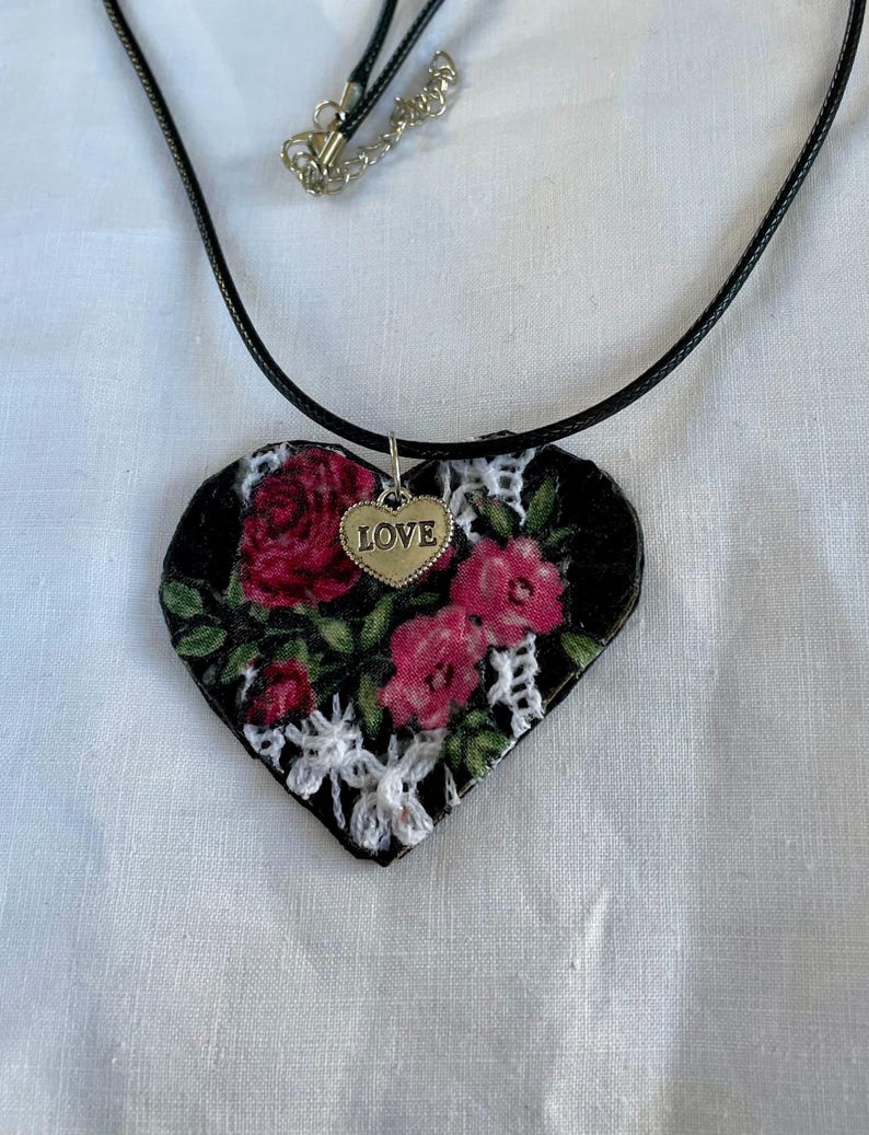 May include: A heart-shaped pendant necklace with a black cord. The pendant features a floral design with red roses and white lace on a black background. A small silver heart charm with the word "LOVE" hangs below the pendant.