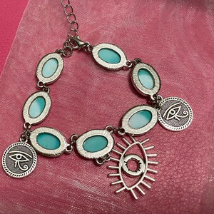 May include: Silver-toned bracelet featuring oval turquoise-colored stones and circular charms with an eye symbol. The bracelet has an adjustable chain and a dangling open-eye pendant. The jewelry is displayed on a pink fabric.