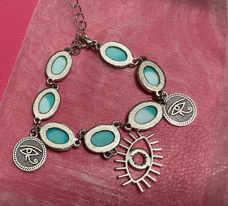 May include: Silver-toned bracelet featuring oval turquoise-colored stones and circular charms with an eye symbol. The bracelet has an adjustable chain and a dangling open-eye pendant. The jewelry is displayed on a pink fabric.