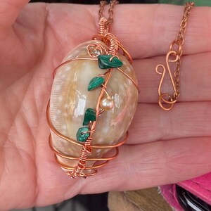 May include: A handcrafted pendant necklace featuring a large, cream-coloured seashell wrapped in copper wire. The shell is adorned with green gemstone leaves and a small pearl. The necklace chain and clasp are also copper.