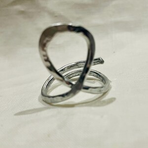 May include: A silver-colored ring with a unique, abstract design. The ring features a looped top and a spiral base, crafted with a hammered texture. The metal reflects light, creating highlights and shadows. The ring is a piece of jewelry.