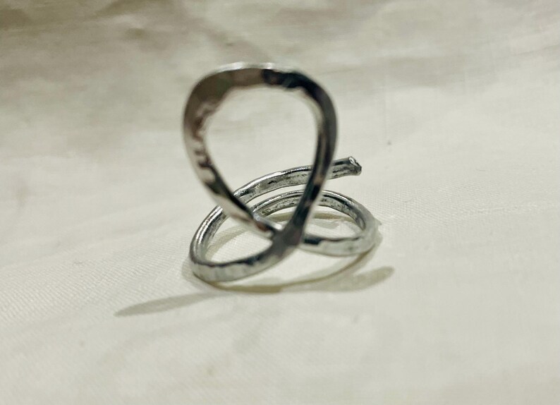 May include: A silver-colored ring with a unique, abstract design. The ring features a looped top and a spiral base, crafted with a hammered texture. The metal reflects light, creating highlights and shadows. The ring is a piece of jewelry.