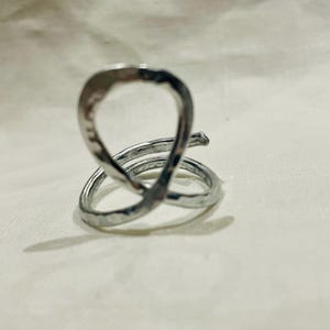 May include: A silver-colored ring with a unique, abstract design. The ring features a looped upper portion and a spiral base, creating an eye-catching, modern aesthetic. The metal has a hammered texture, adding visual interest to the jewelry.