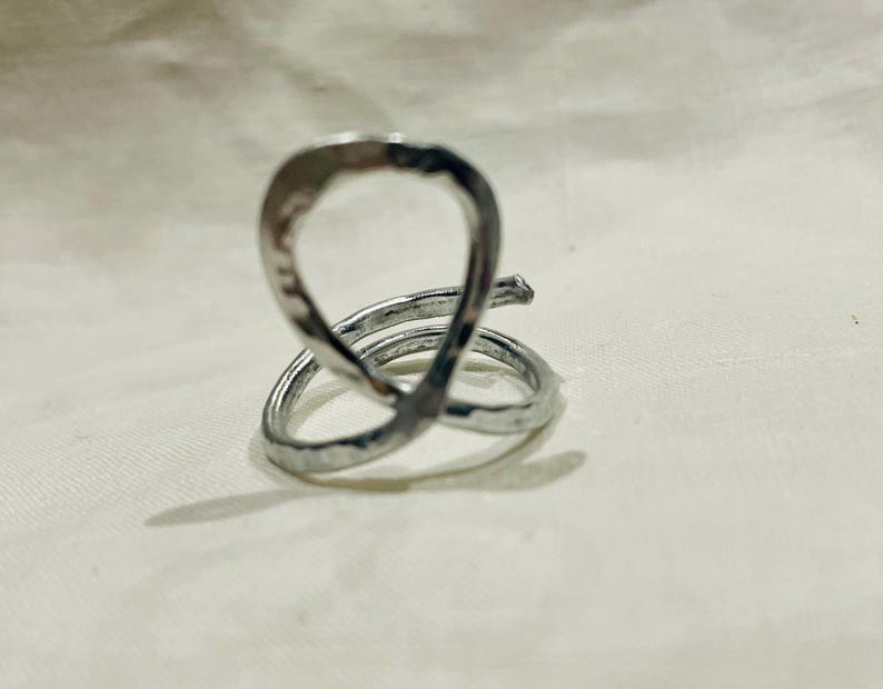 May include: A silver-colored ring with a unique, abstract design. The ring features a looped upper portion and a spiral base, creating an eye-catching, modern aesthetic. The metal has a hammered texture, adding visual interest to the jewelry.