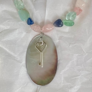 May include: A handmade necklace with a silver key pendant and a strand of colourful gemstone beads. The beads are in shades of pink, green, and blue. The oval pendant has a pearlescent finish. The necklace is a unique piece of jewellery.