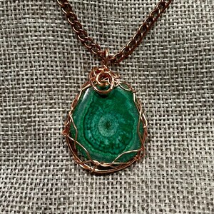 Peut inclure: Un collier avec une chaîne de couleur cuivre et un pendentif. Le pendentif présente une pierre verte polie en forme de goutte d'eau, finement enveloppée dans du fil de cuivre. La pierre a un motif tourbillonnant. Le collier est présenté sur une surface grise texturée.