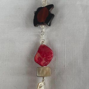 May include: A silver chain necklace with a pendant featuring a black and brown stone, a red stone, a beige rectangular stone, and a white stone. The stones are connected by silver wire.