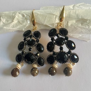 May include: A pair of dangle earrings featuring clusters of faceted black oval stones set in dark metal frames. Each earring hangs from a gold-tone hook and terminates in two small, round, brown-toned beads suspended by light-colored spacers.