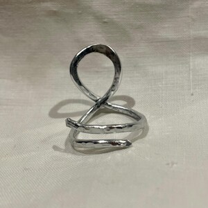 May include: A silver-colored ring with an abstract design. The ring features a looped top and a spiral base, crafted from a hammered metal. The ring is displayed on a white surface.