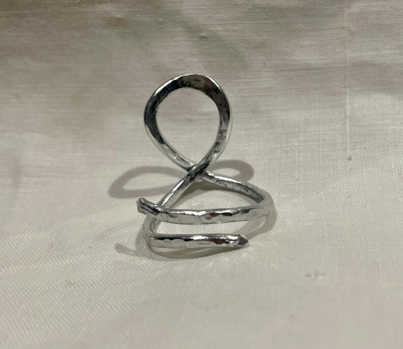 May include: A silver-colored ring with an abstract design. The ring features a looped top and a spiral base, crafted from a hammered metal. The ring is displayed on a white surface.
