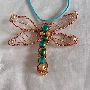 以下が含まれることがあります： 銅線で作られたトンボのペンダント。ターコイズとクリーム色のビーズが使用されています。トンボの羽は銅線で複雑に織られており、体にはビーズが飾られています。ライトブルーのスエードコードから吊り下げられています。